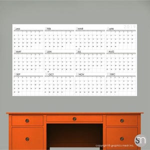 Puede incluir: Un gran calendario blanco de 2019 con cuadrículas mensuales está montado en una pared gris. El calendario está encima de un escritorio de madera naranja con cajones y un espacio central abierto. El calendario muestra los meses de enero a diciembre.