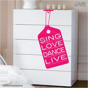 May include: A white chest of drawers with a pink tag-shaped sticker. The sticker displays the words "SING, LOVE, DANCE, LIVE" in white, accompanied by motivational phrases. The chest has five drawers and a simple design.