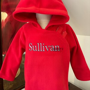May include: A bright red fleece hooded pullover featuring long sleeves and an asymmetrical front closure with hook-and-loop fasteners. The name Sullivan is embroidered in white lettering with a dark outline across the center of the chest area.