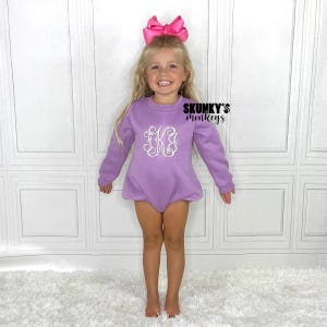 May include: A young child wearing a lavender romper with a white monogram and a large pink bow. The romper has long sleeves and a round neckline. The text "SKUNKY'S monkeys" is visible on the right side.