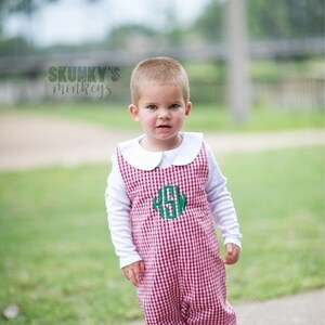 May include: A young boy wearing a white long-sleeve shirt and red and white gingham overalls with a green monogram. The overalls have a white Peter Pan collar.