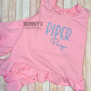 May include: Pink sleeveless shirt with a ruffled neckline and hem. The shirt has the text "Skunky's Monkeys" and "Piper Paige" embroidered in blue.