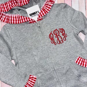 May include: A gray zip-up hoodie with red and white gingham trim on the hood, cuffs, and hem. The hoodie features a red monogram on the chest and a tag that reads "SKUNKY'S monkeys".