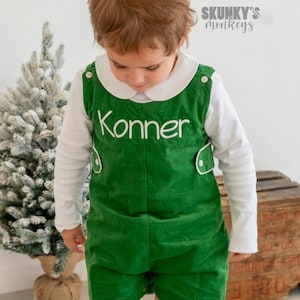 May include: A young child wearing a green corduroy overall with the name "Konner" embroidered on the front. The overall has white piping and buttons. The child is also wearing a white long-sleeved shirt with a white collar. The child is standing in front of a white wall with a small artificial Christmas tree to the left.
