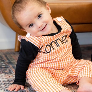 May include: A baby boy wearing a black long-sleeved shirt and orange and white gingham overalls. The overalls have the name "Konner" embroidered on the front. The baby is sitting on a patterned rug.