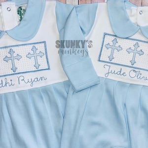 May include: Two light blue baby onesies with white collars and embroidered crosses. The onesies have the names "Bodhi Ryan" and "Jude Oliver" embroidered on them.