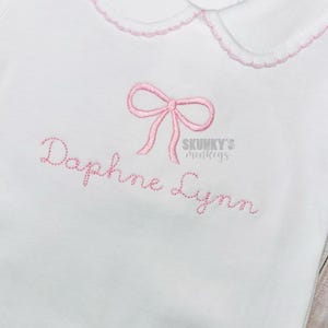 May include: White baby onesie with a pink bow and the name "Daphne Lynn" embroidered on the front. The onesie has a pink and white scalloped collar.