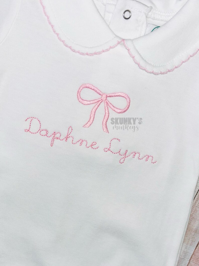 May include: White baby onesie with a pink bow and the name "Daphne Lynn" embroidered on the front. The onesie has a pink and white scalloped collar.