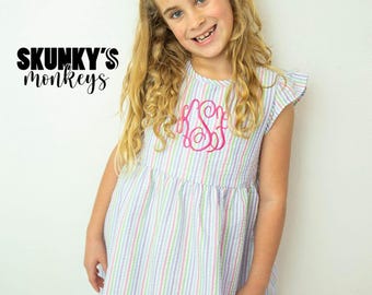 May include: A young girl wearing a white and rainbow striped dress with a pink bow in her hair. The dress has a pink embroidered monogram on the front. The text "SKUNKY'S monkeys" is visible in the image.