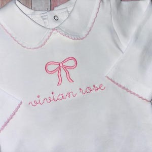 May include: White long-sleeve baby shirt with a pink embroidered bow and the name "vivian rose" in pink thread.