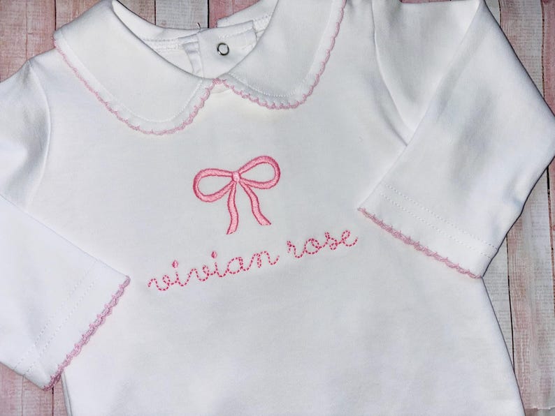 May include: White long-sleeve baby shirt with a pink embroidered bow and the name "vivian rose" in pink thread.