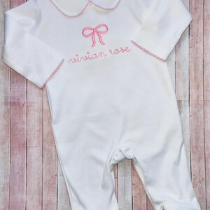 May include: White baby onesie with a pink bow and the name "Vivian Rose" embroidered on the front.