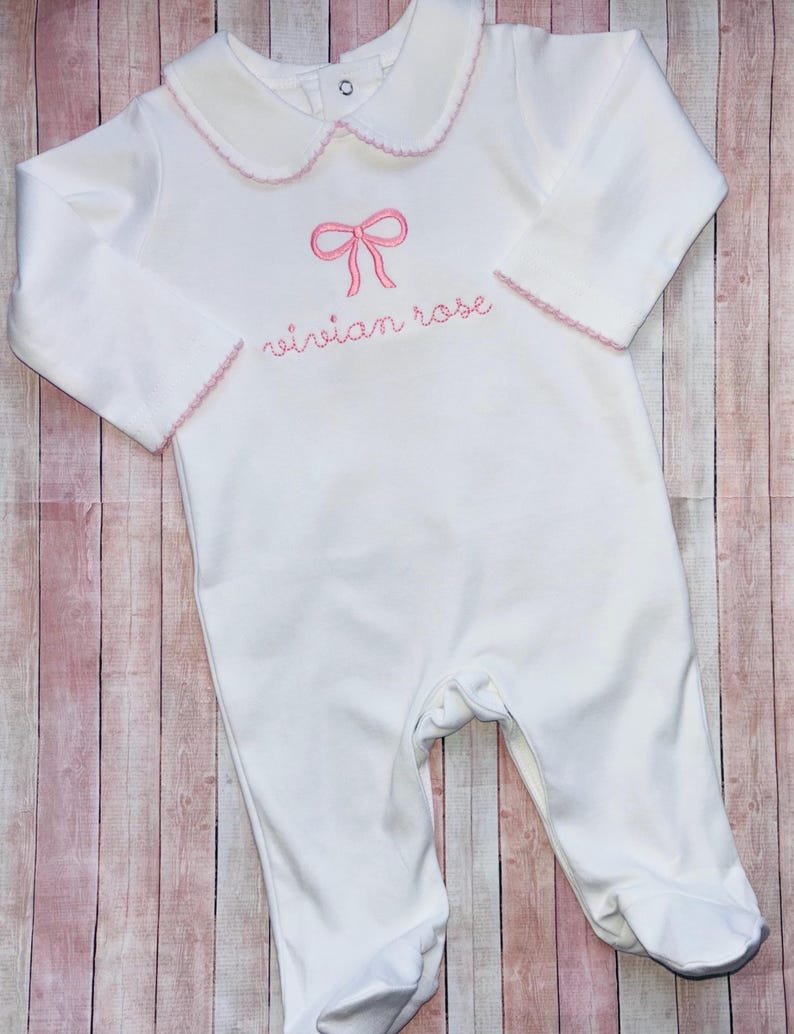 May include: White baby onesie with a pink bow and the name "Vivian Rose" embroidered on the front.