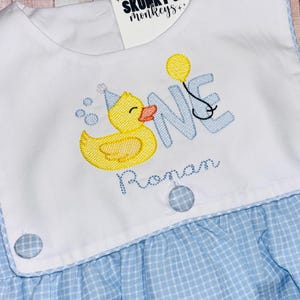 May include: A white and light blue gingham dress featuring an embroidered yellow rubber duck wearing a blue party hat, the word ONE in light blue, a yellow balloon, and the name Ronan in blue script, with two blue gingham buttons at the waist.