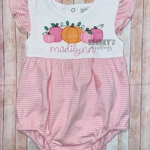 May include: Pink and white striped baby onesie with a white ruffle on the sleeves. The onesie has an embroidered design of pumpkins with vines and the name "madilynn" in cursive.