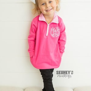 May include: A pink fleece pullover with a white gingham collar and a white monogram on the front. The pullover is worn by a young girl with blonde hair and a white bow. The girl is standing on a white couch. The text "SKUNKY'S monkeys" is visible in the bottom right corner of the image.