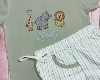 May include: A sage green short-sleeve t-shirt featuring embroidered giraffe, elephant, and lion figures above the name HENRY in light blue thread. The shirt includes a Skunky's Monkeys brand tag. Paired with white shorts featuring thin green stripes.