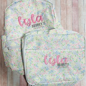 May include: A floral-patterned backpack and matching lunch bag set. The bags feature a pastel floral print with pink embroidered text that reads "Isla" and the text "SKUNKY'S monkeys". Both bags have zippers and are on a wooden background.