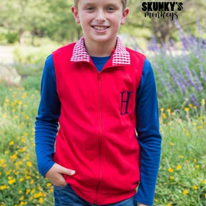 May include: A young boy wearing a red zippered vest with a white and red checkered collar, a blue long-sleeved shirt, blue jeans, and the text "SKUNKY'S monkeys" on the right side of the image.