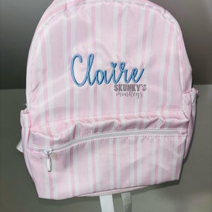 May include: Pink and white striped backpack with the name "Claire" embroidered in blue script. The backpack has a front zippered pocket, gray straps, and the text "SKUNKY'S monkeys" in gray.