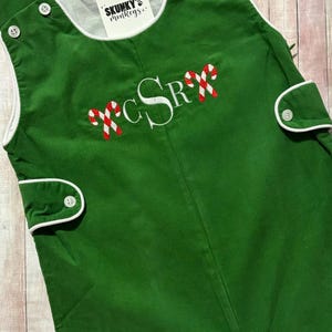 May include: Green corduroy Christmas-themed romper with white trim and buttons. Features embroidered initials and candy cane accents. The tag reads "Skunky's Monkeys".