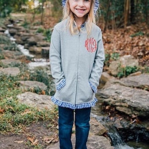 May include: A young girl wearing a gray zip-up hoodie with a red monogram, blue jeans, and white sandals. The hoodie has blue and white gingham trim on the hood, pockets, and hem. She is standing near a stream.