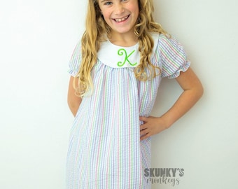 May include: A young girl wearing a white and pink striped dress with a white Peter Pan collar and a green embroidered letter "K". She is smiling and has a pink bow in her hair. The text "SKUNKY'S monkeys" is visible in the bottom right corner of the image.
