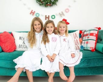 May include: Three young people in white nightgowns with personalized monograms and red trim sit on a teal velvet couch. Decorative pillows and a Christmas garland with a "Ho Ho Ho" banner are in the background.