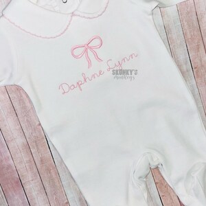 May include: White baby onesie with a pink bow and the name "Daphne Lynn" embroidered on the front. The onesie has a scalloped collar and is made of soft cotton.