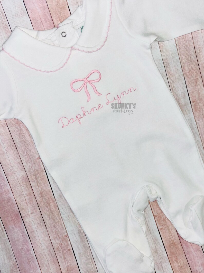 May include: White baby onesie with a pink bow and the name "Daphne Lynn" embroidered on the front. The onesie has a scalloped collar and is made of soft cotton.