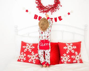 May include: A child in Christmas pajamas with the name "Quinn" on the back, reaching for a red berry wreath. Red snowflake pillows and a "BELIEVE" banner decorate the white wall, creating a festive holiday scene.