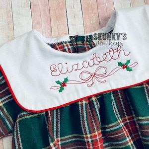 May include: A green and red plaid dress with a white collar and red trim. The collar has the name "Elizabeth" embroidered on it with a red bow and holly.