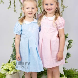 May include: Two young girls wearing matching dresses with their names embroidered on the front. The girl on the left is wearing a light blue dress with the name "Kylie" embroidered on it. The girl on the right is wearing a pink dress with the name "Shiloh" embroidered on it. Both girls are wearing white collars and have their hair styled with bows.