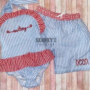 May include: A blue and white striped swimsuit and matching shorts set. The swimsuit has red gingham trim and the name "audrey" embroidered in red. The shorts have the name "dean" embroidered in red.