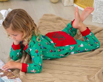 May include: A child wearing green pajamas with a festive print of toy soldiers and fairies, accented with red cuffs and a red back panel. The child is lying on a tan blanket and reading a book.