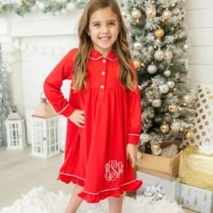 May include: A red long-sleeved nightgown with white trim and a monogram on the front. The girl is standing on a white fluffy rug in front of a Christmas tree.