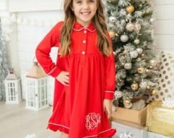 May include: A red long-sleeved nightgown with white trim and a monogram on the front. The girl is standing on a white fluffy rug in front of a Christmas tree.