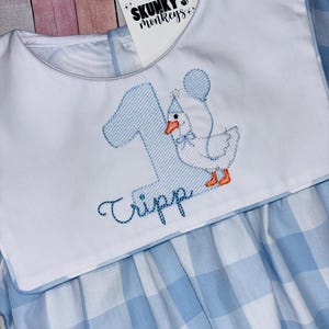 May include: A baby's blue and white gingham dress with a white bib featuring a blue embroidered number 1, a cartoon goose with a party hat and balloon, and the name "Tripp". The dress has a Peter Pan collar.