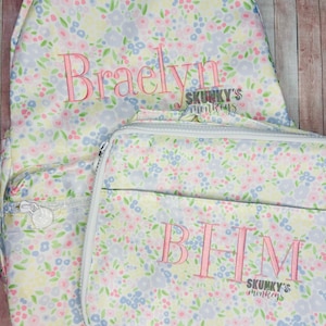 May include: A floral-patterned backpack and matching lunch bag. The backpack has the name "Braelyn" embroidered in pink. The lunch bag has the initials "BHM" embroidered in pink. Both items feature a white zipper and the text "SKUNKY'S monkeys".