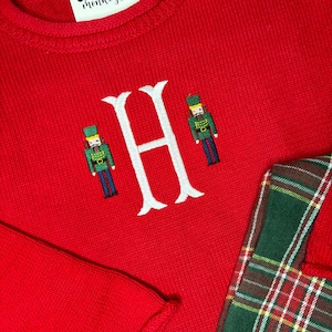 May include: A red knit sweater with a white embroidered letter "H" and two toy soldier figures. The sweater has a crew neck and a tag that reads "Skunky's Monkeys +." A green and red plaid pattern is visible on the right side.