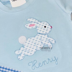 May include: Light blue t-shirt with a white bunny applique featuring a blue and white pattern. The bunny has pink accents and is jumping. The name "Henry" is embroidered in blue. The shirt has a blue and white gingham pattern at the bottom.