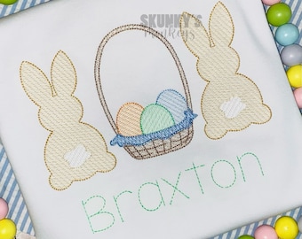 May include: A white t-shirt with an embroidered design of two bunnies and a basket of Easter eggs. The name "Braxton" is embroidered below the design.