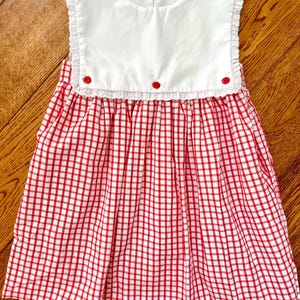 May include: A red and white checkered dress with a white collar. The collar is trimmed with lace and has three red buttons. The dress is sleeveless and has a gathered skirt. The dress is made of a lightweight fabric.