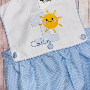 May include: A light blue and white gingham bubble romper featuring a white bib front with an embroidered light blue number one, a yellow sun with a smiling face, and the name Colin in dark blue script. Three gingham buttons accent the bib.