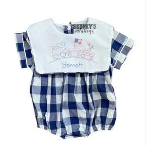 May include: A blue and white gingham baby romper with a white bib collar. The collar features an embroidered train, flag, and fireworks design, with the name "Bennett" below. The romper has short sleeves and a round neckline.