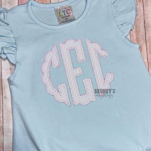 May include: Light blue baby romper with ruffled sleeves and a round neckline. The romper features a monogram in white with pink polka dots and a scalloped edge. The tag reads "LTC Made in China".
