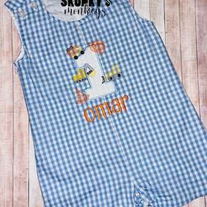May include: Blue and white gingham romper with the number "1" and construction vehicle appliqués. The name "Omar" is embroidered in orange. The romper has button closures on the straps and at the bottom. The text "SKUNKY'S monkeys" is at the top.