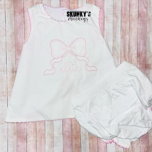 May include: White baby dress and matching bloomers with pink trim. The dress features a pink embroidered bow and the name "Ellie". The text "Skunky's Monkeys" is at the top. The bloomers have an elastic waistband.