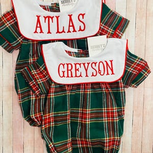 May include: Two green, red, and yellow plaid baby onesies with white sailor collars and red trim. The onesies are personalized with the names "Atlas" and "Greyson" in red embroidery.