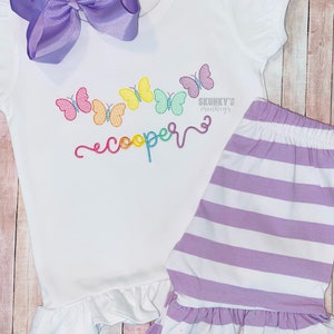 May include: A white baby outfit with a rainbow butterfly design and the name "cooper" embroidered on the front. The outfit includes a short-sleeved shirt with a ruffled hem and a pair of striped shorts with a ruffled hem.